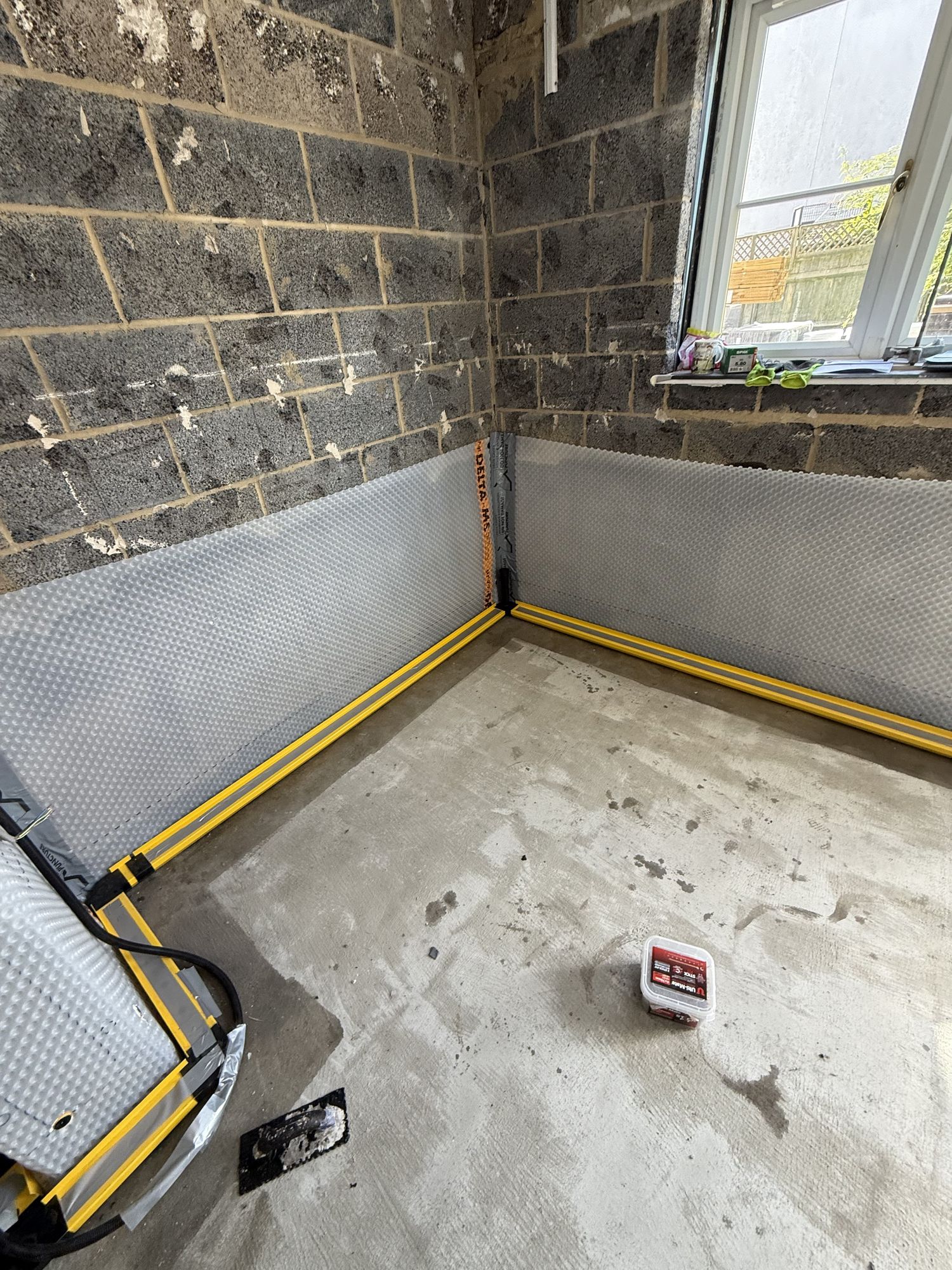 Finished concrete slab with two coats of waterproof resin applied — yellow drain pipes running to the sump visible, Delta-MS membrane fixed to walls